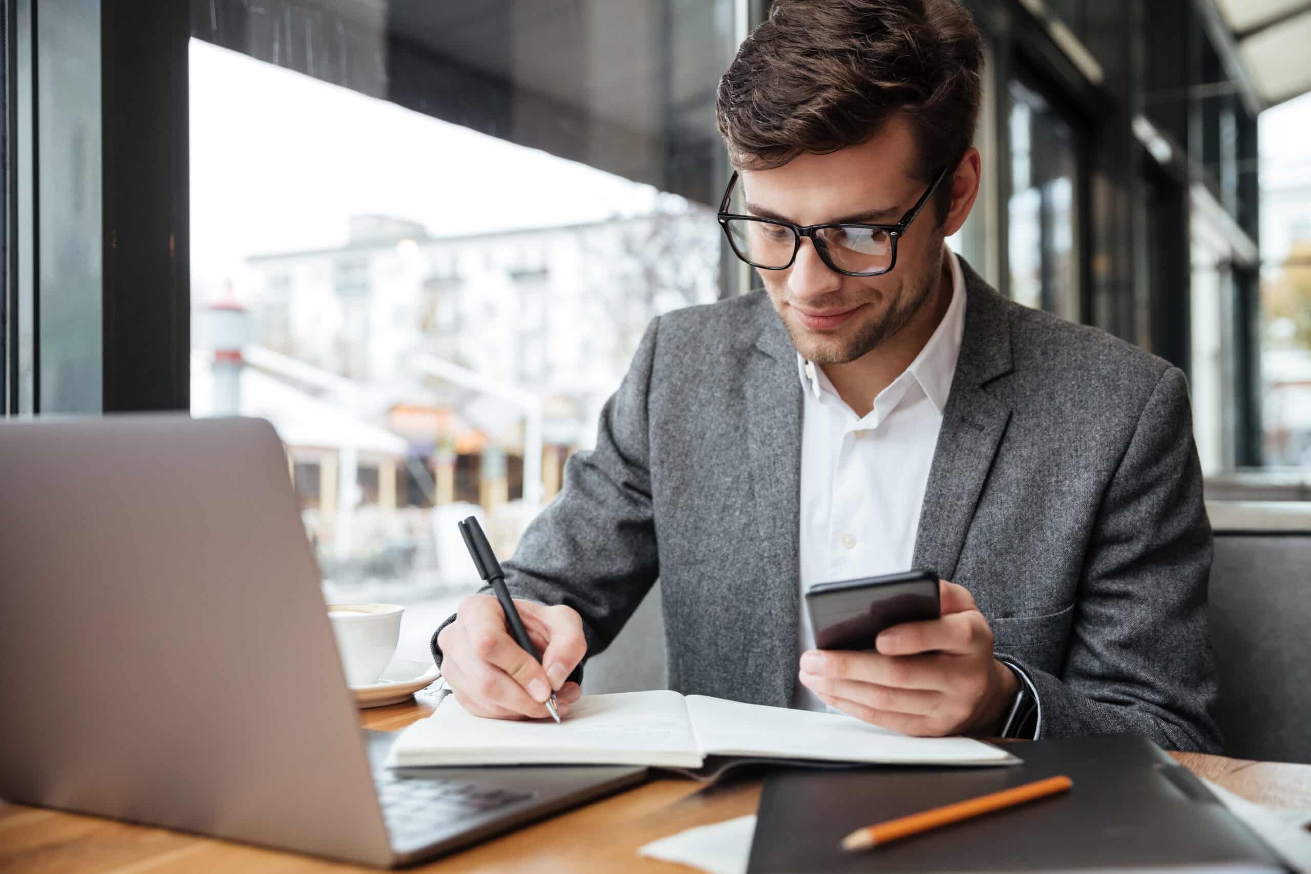 smiling-businessman-eyeglasses-sitting-by-table-cafe-with-laptop-computer-while-using-smartphone-writing-something-scaled.jpg